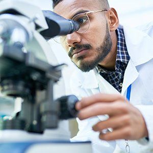 A researcher in a white lab coat uses a microscope, looking through the eyepiece while adjusting the focus knob.