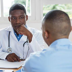 A doctor in a white coat and stethoscope listens to a patient during a consultation in a bright, modern office.