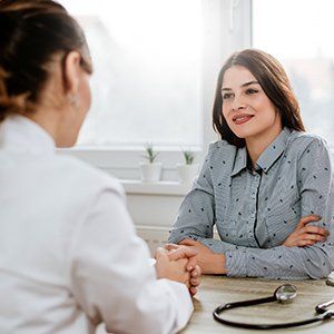 A professional in a white coat sits across from a patient, both engaging in a calm conversation at a desk with a stethoscope.