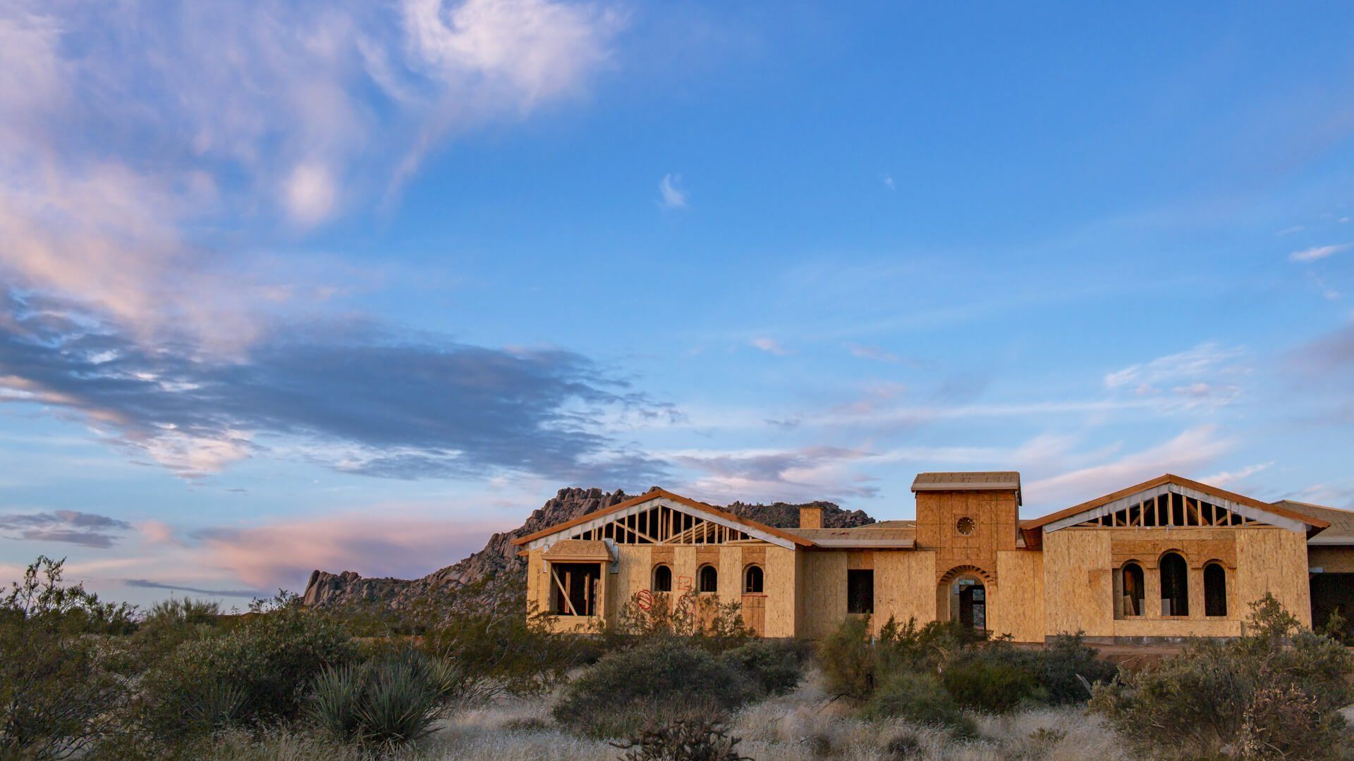 An unfinished tan stucco house stands in a desert landscape against a blue and pink twilight sky.