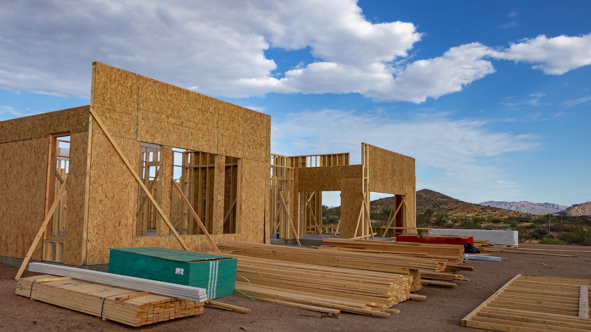 Unfinished house frame under a bright blue sky with scattered clouds and desert hills in the background.