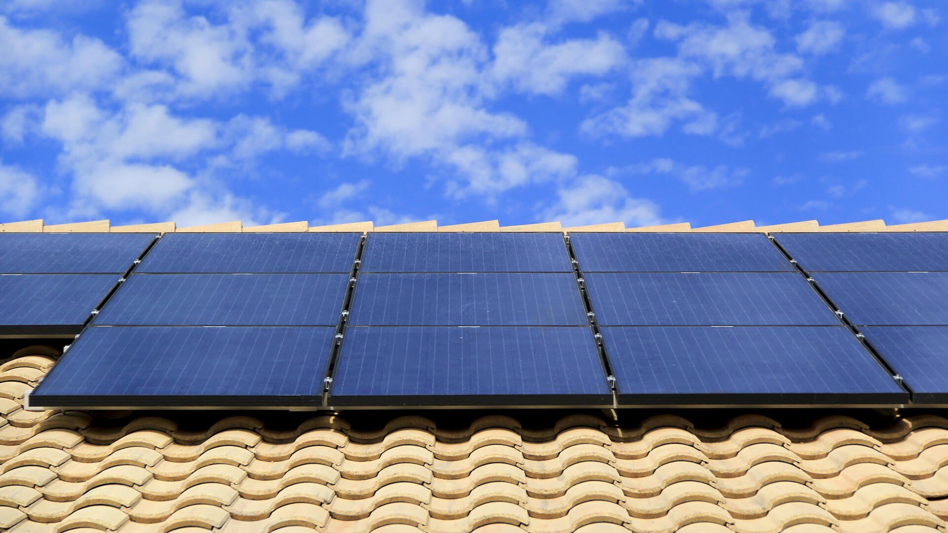 Dark blue solar panels installed on a textured, light-colored tile roof against a bright blue sky with white clouds.