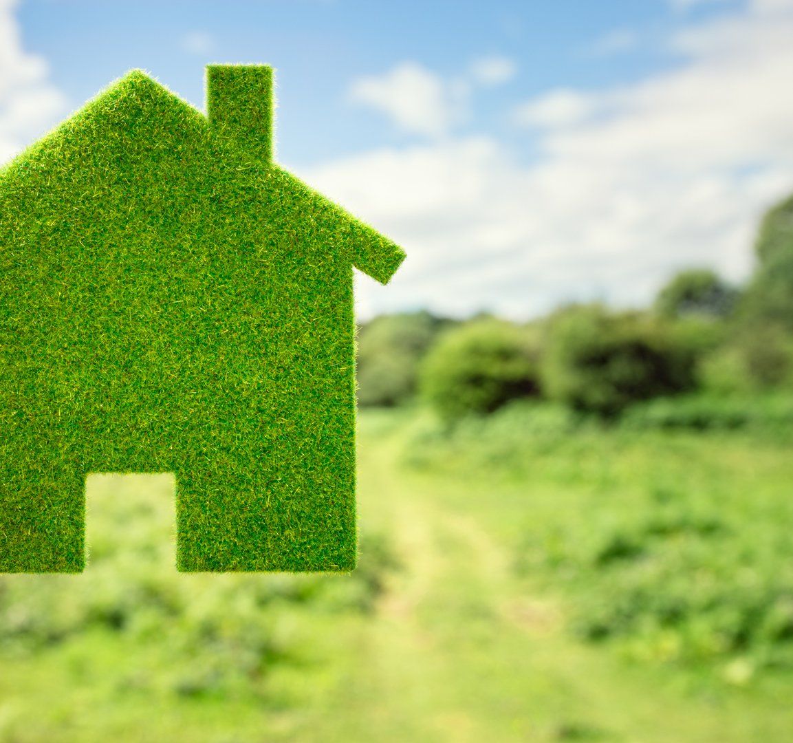 A small, green house-shaped cutout held up against a blurry background of a green field and a blue sky.