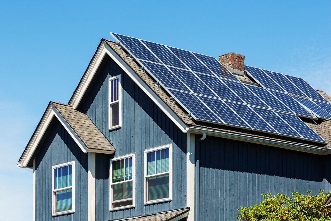A blue house with white trim features a roof covered in dark solar panels against a clear blue sky.