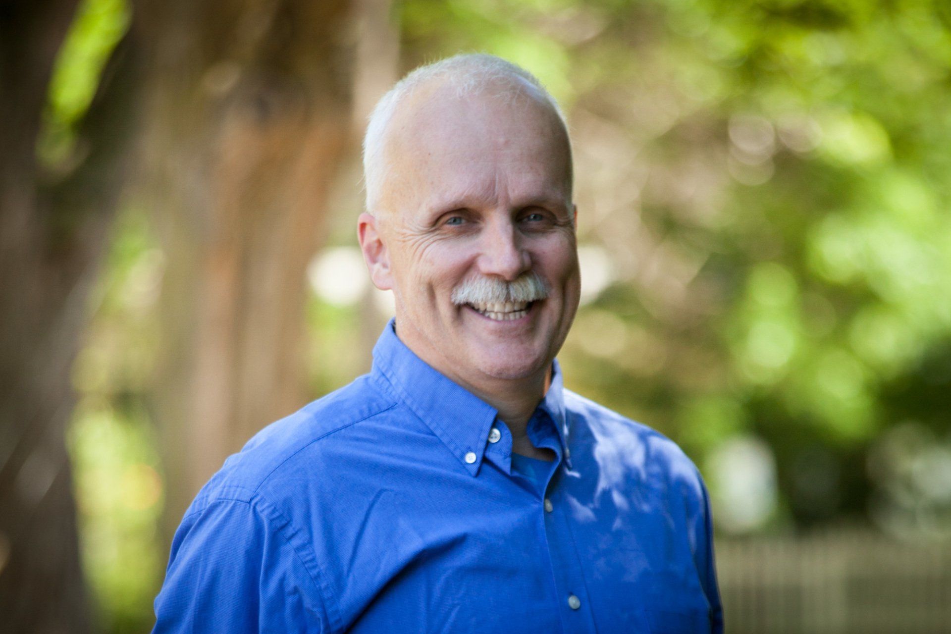 A man with a mustache is wearing a blue shirt and smiling.