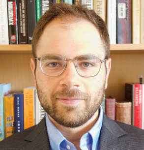 A man wearing glasses and a suit is standing in front of a bookshelf with books on it including one called mindmix