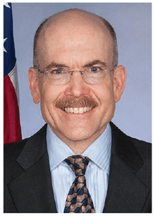 A man in a suit and tie is smiling in front of an american flag.