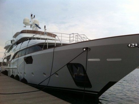 Un grande yacht di lusso bianco ormeggiato in un porto turistico, visto dalla prua su acque calme.