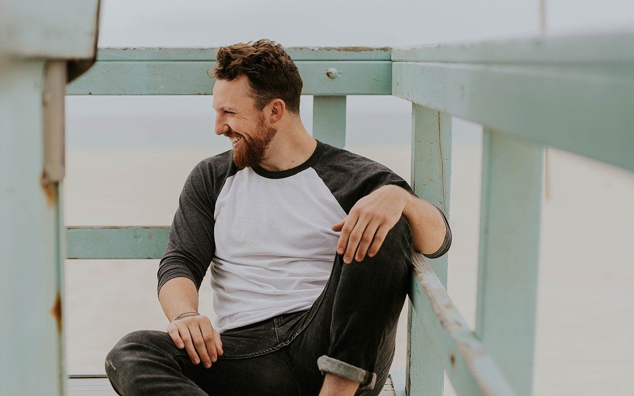 A man with a beard is sitting on a railing on a beach.