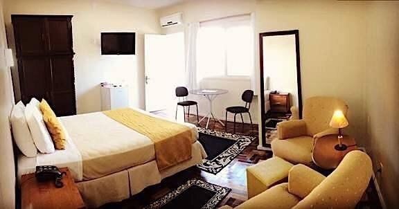 Quarto luminoso com cama, poltrona, espelho de corpo inteiro e abajur junto à janela.