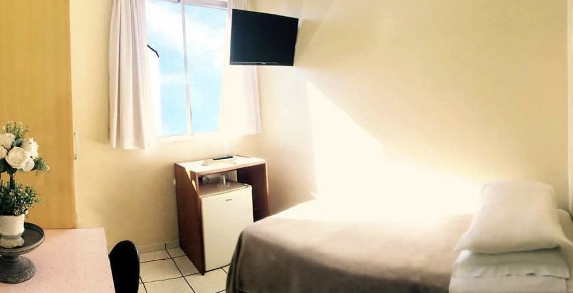 Quarto pequeno com cama, TV fixada na parede, janela e escrivaninha junto à janela.