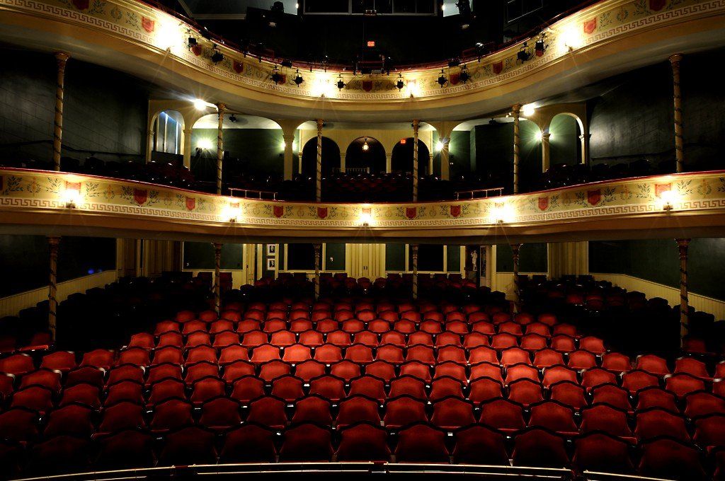 Empty theater with red seats and ornate balconies, viewed from the stage.