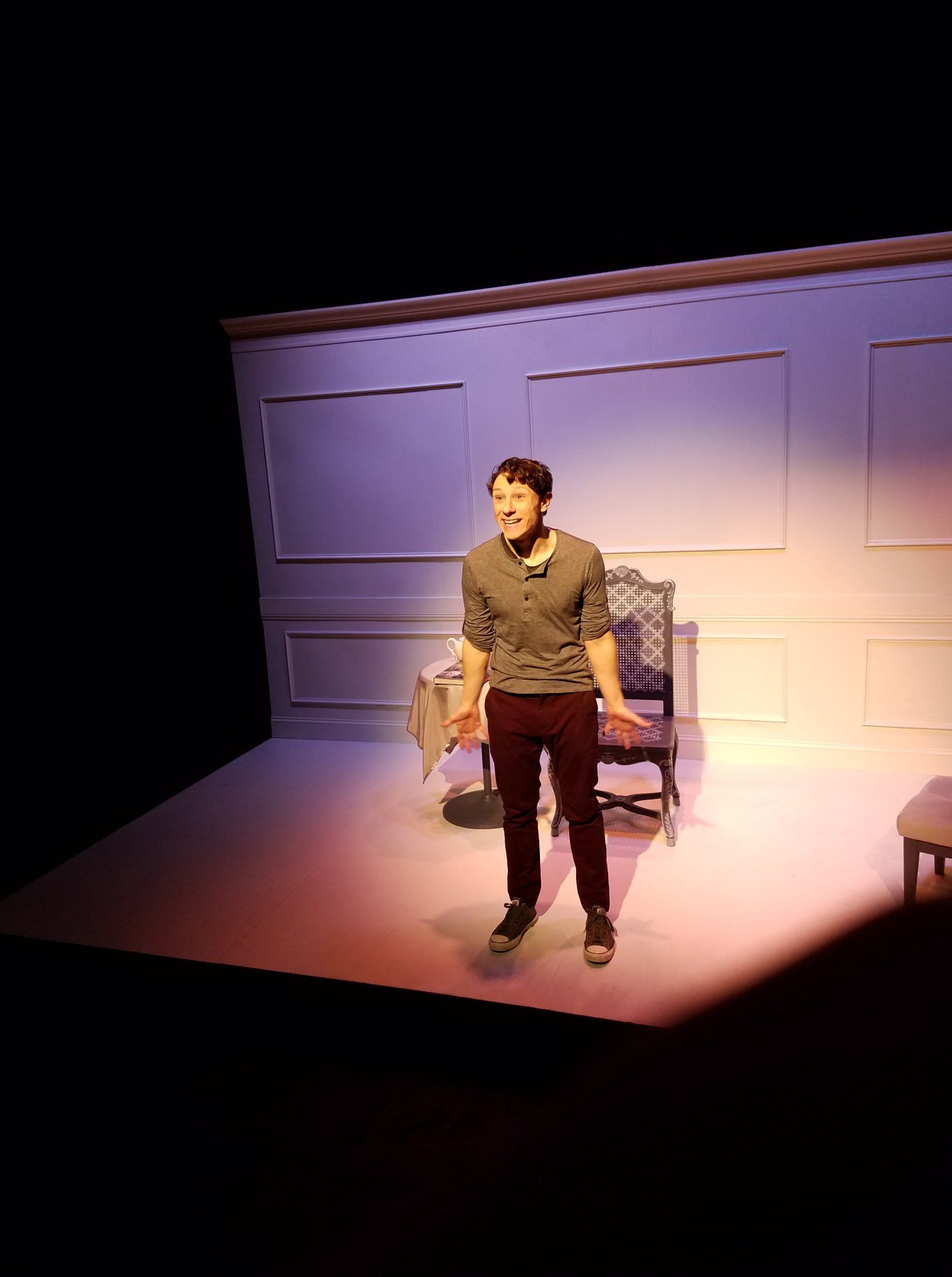 Man standing on a lit stage beside a chair, in front of paneled walls