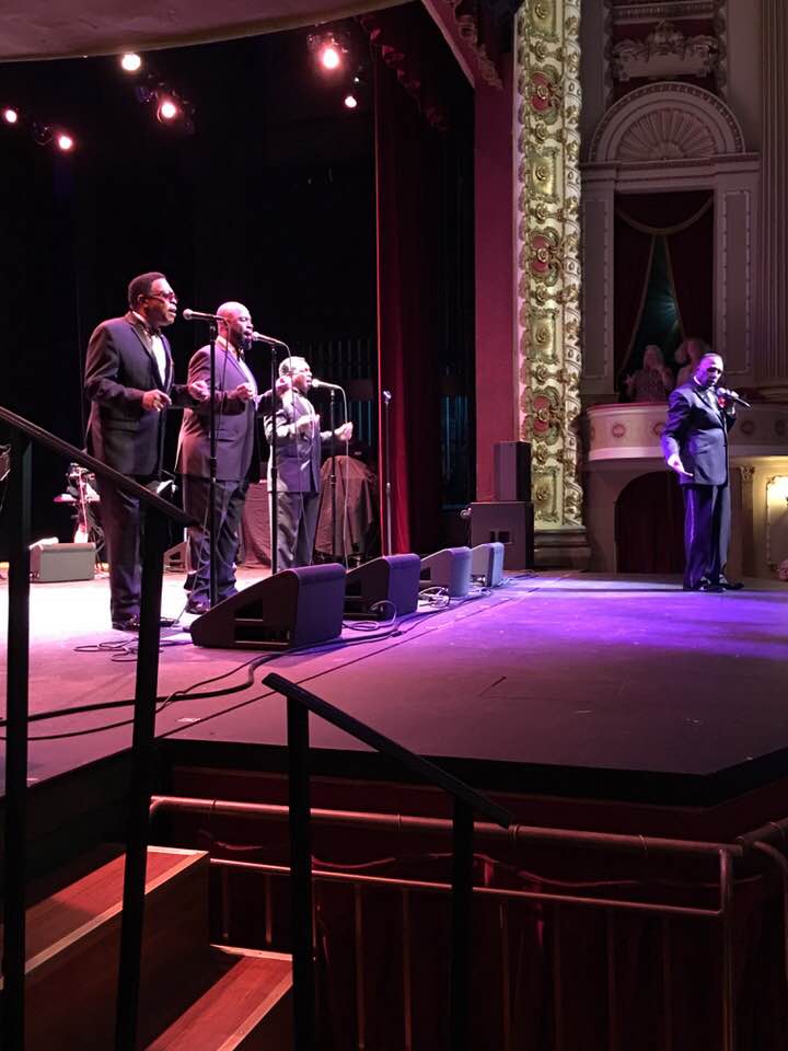 Four performers sing on a stage under pink lights in an ornate theater.