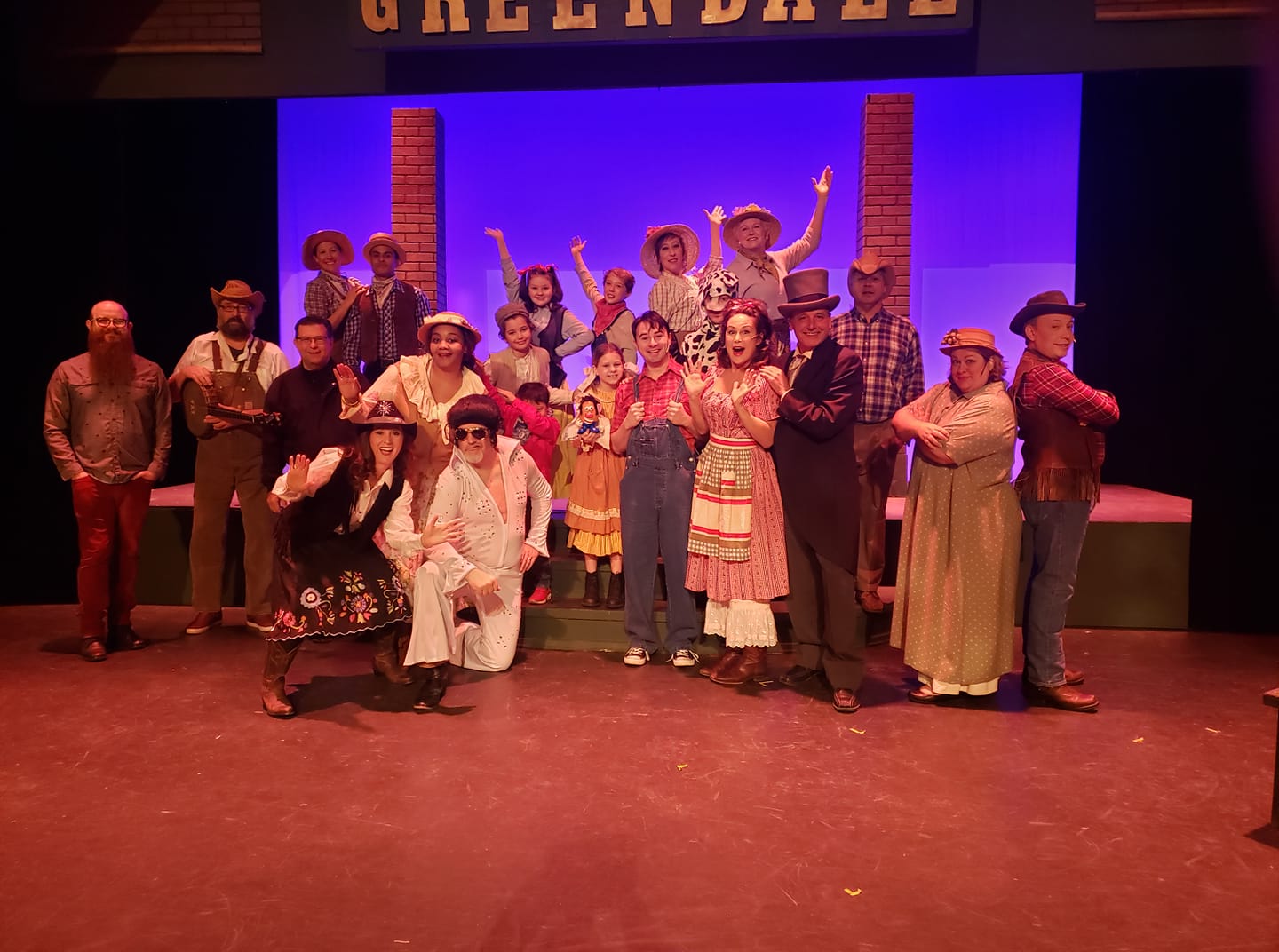 Cast of a stage musical posing under a “Greendale” sign, wearing colorful costumes under purple lighting.