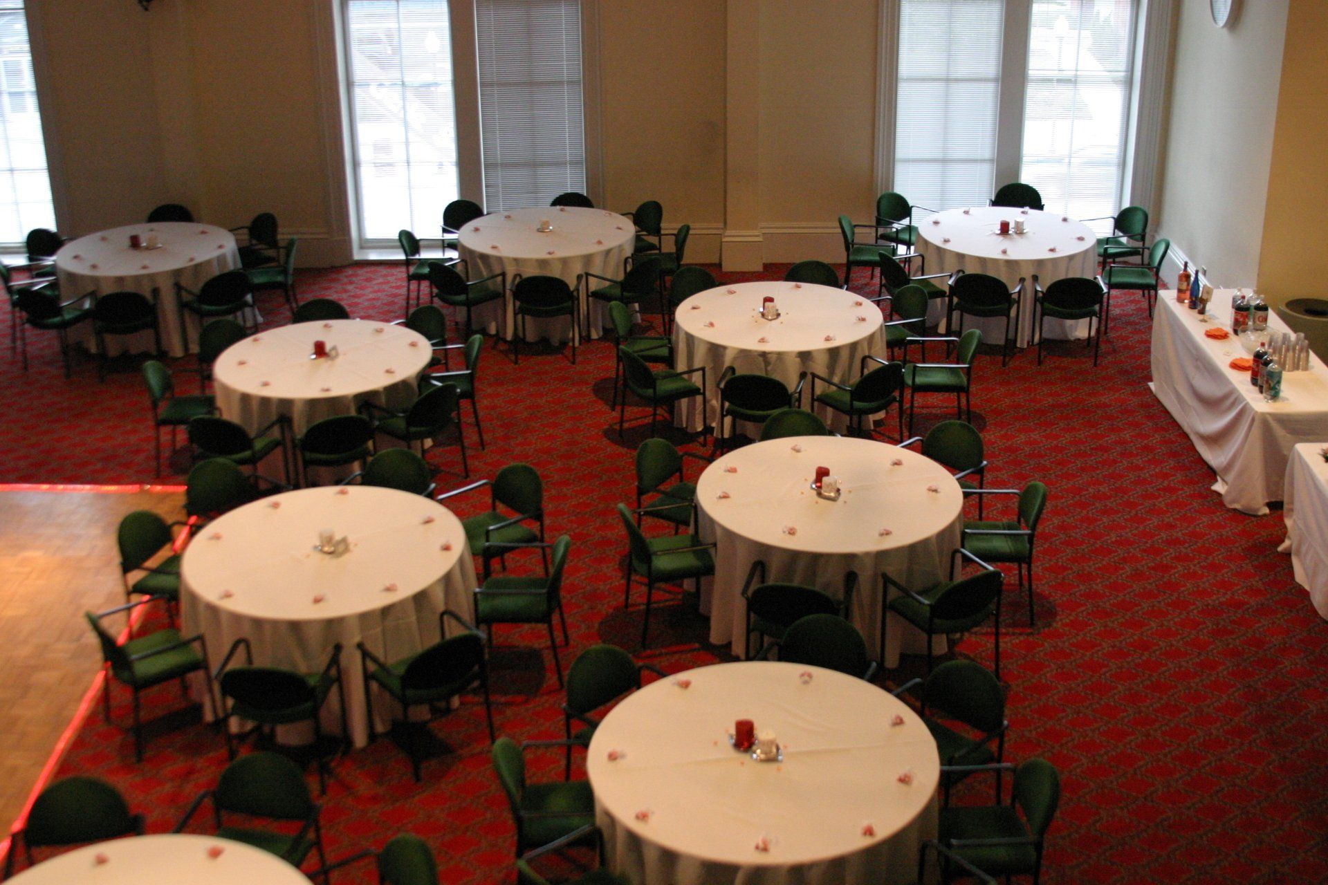 Dining room with round tables and green chairs on a red carpet, set for a banquet near large windows.