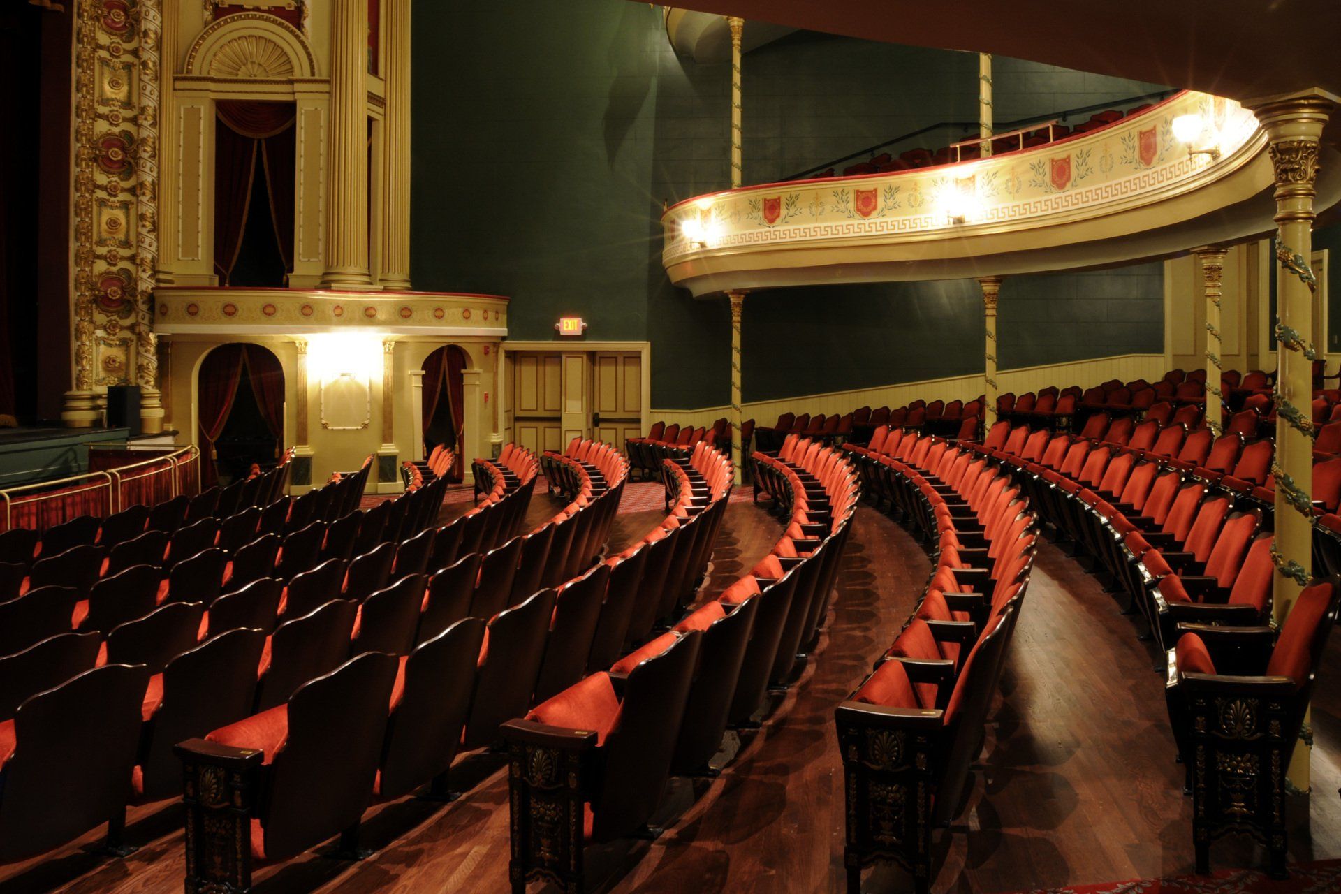Empty red theater seats in a historic ornate auditorium with a lit balcony.