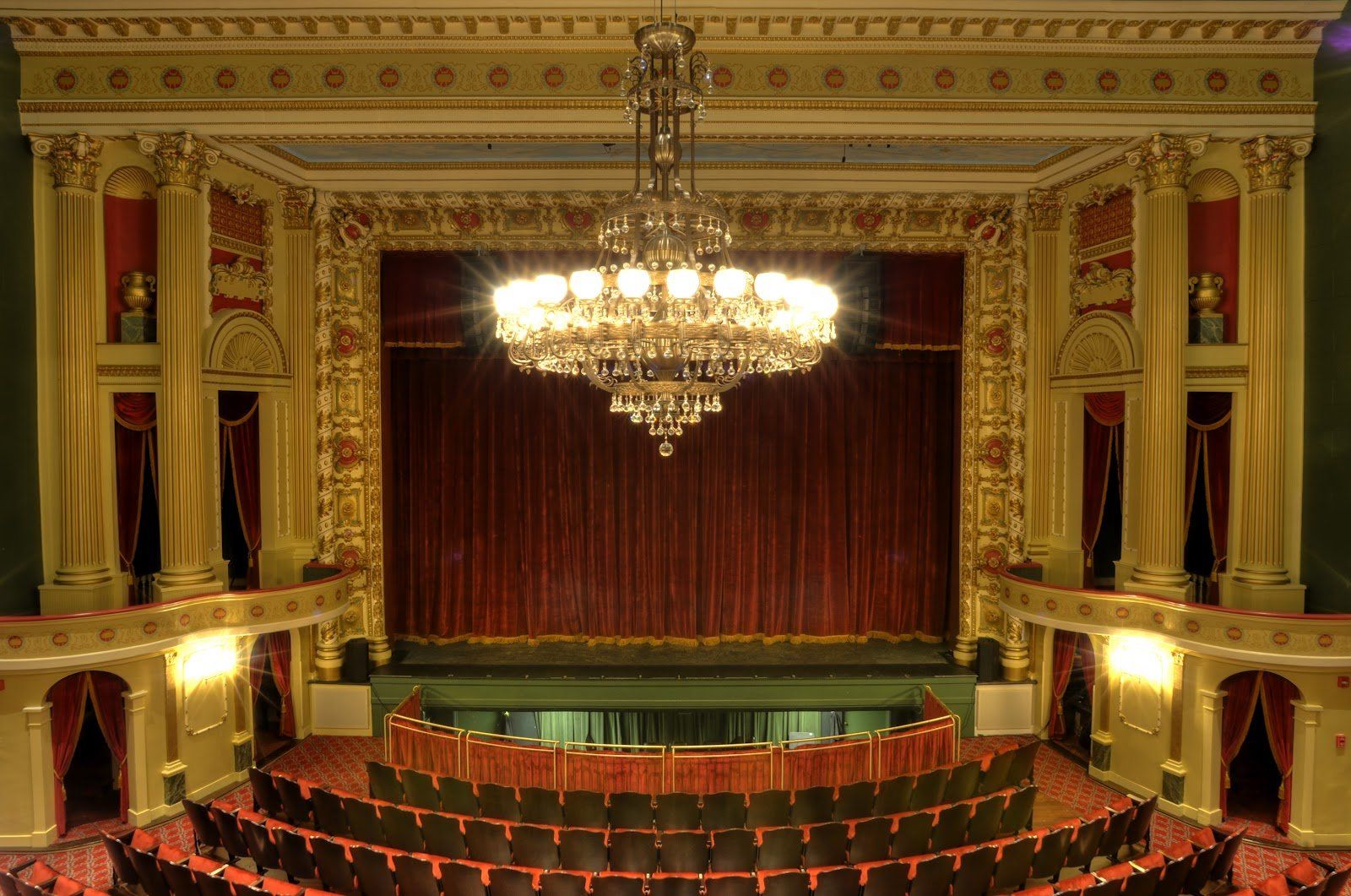 Ornate theater auditorium with red seats, gold decor, and a large chandelier above the stage