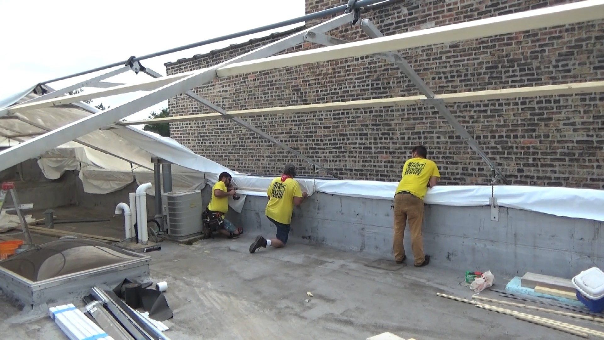 Workers installing construction grade 12mm shrinkwrap along a rooftop wall for construction tent