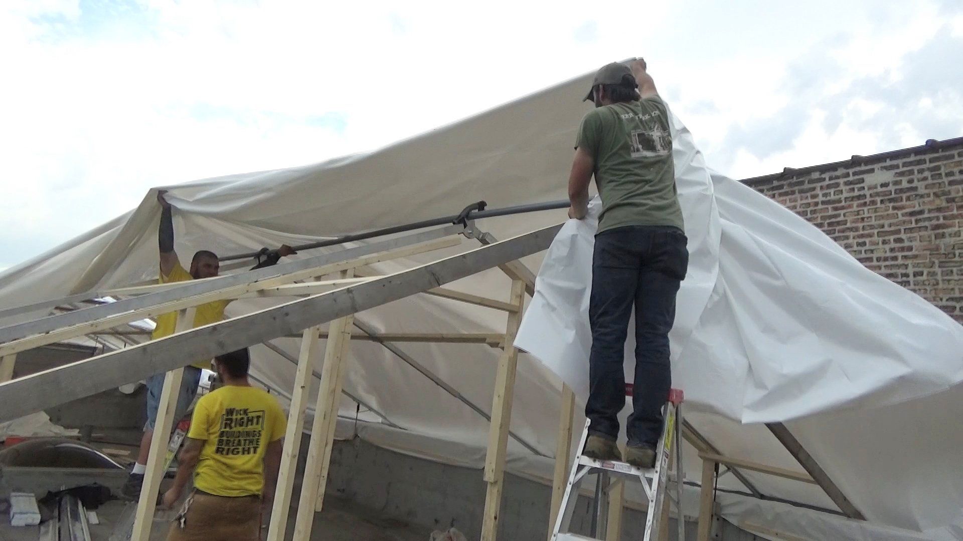 Two workers securing construction grade 12mm shrinkwrap over steel framing for construction tent