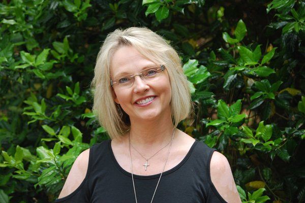 Blonde woman with glasses smiles in front of green bushes, wearing a black top and a necklace.