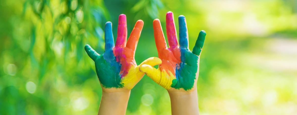 Hands with rainbow-colored paint raised in front of a blurred green background.