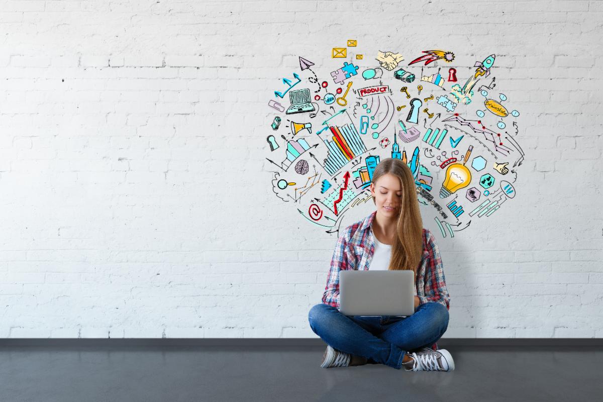Woman using laptop, sitting on floor against white brick wall with colorful brainstorm drawings.