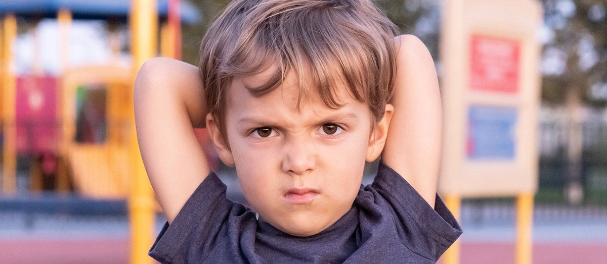 A child with an angry expression, hands behind their head, at a playground.
