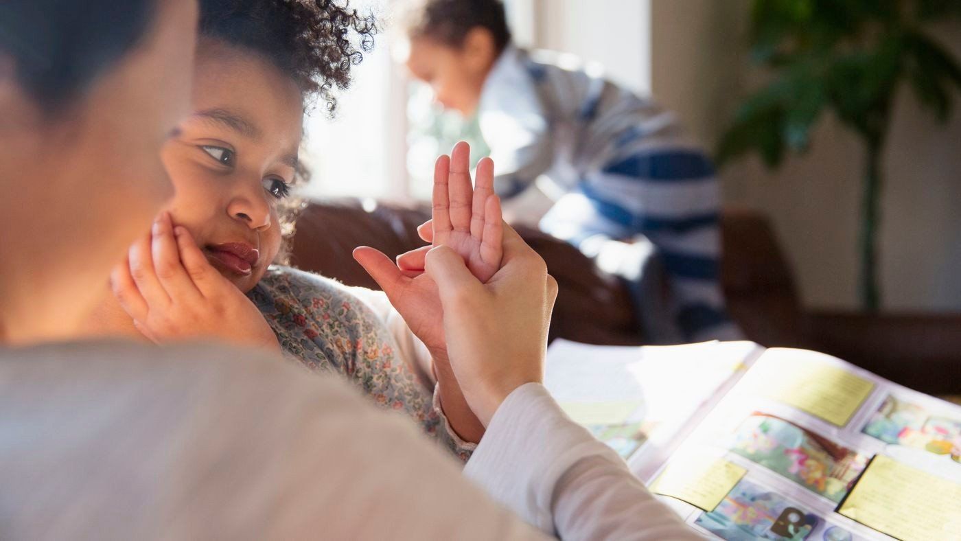 Woman using sign language with a child, reading a book. Another child in the background.