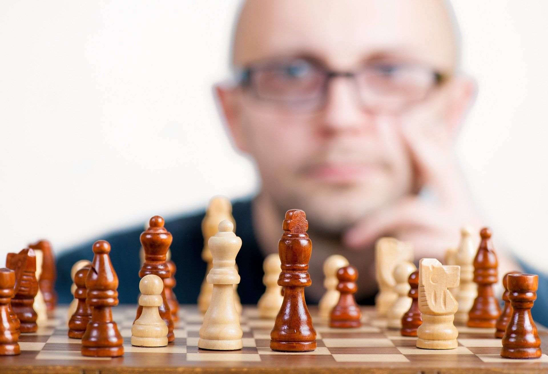 Man with glasses looking at a chessboard, resting chin on hand. Chess pieces in focus.