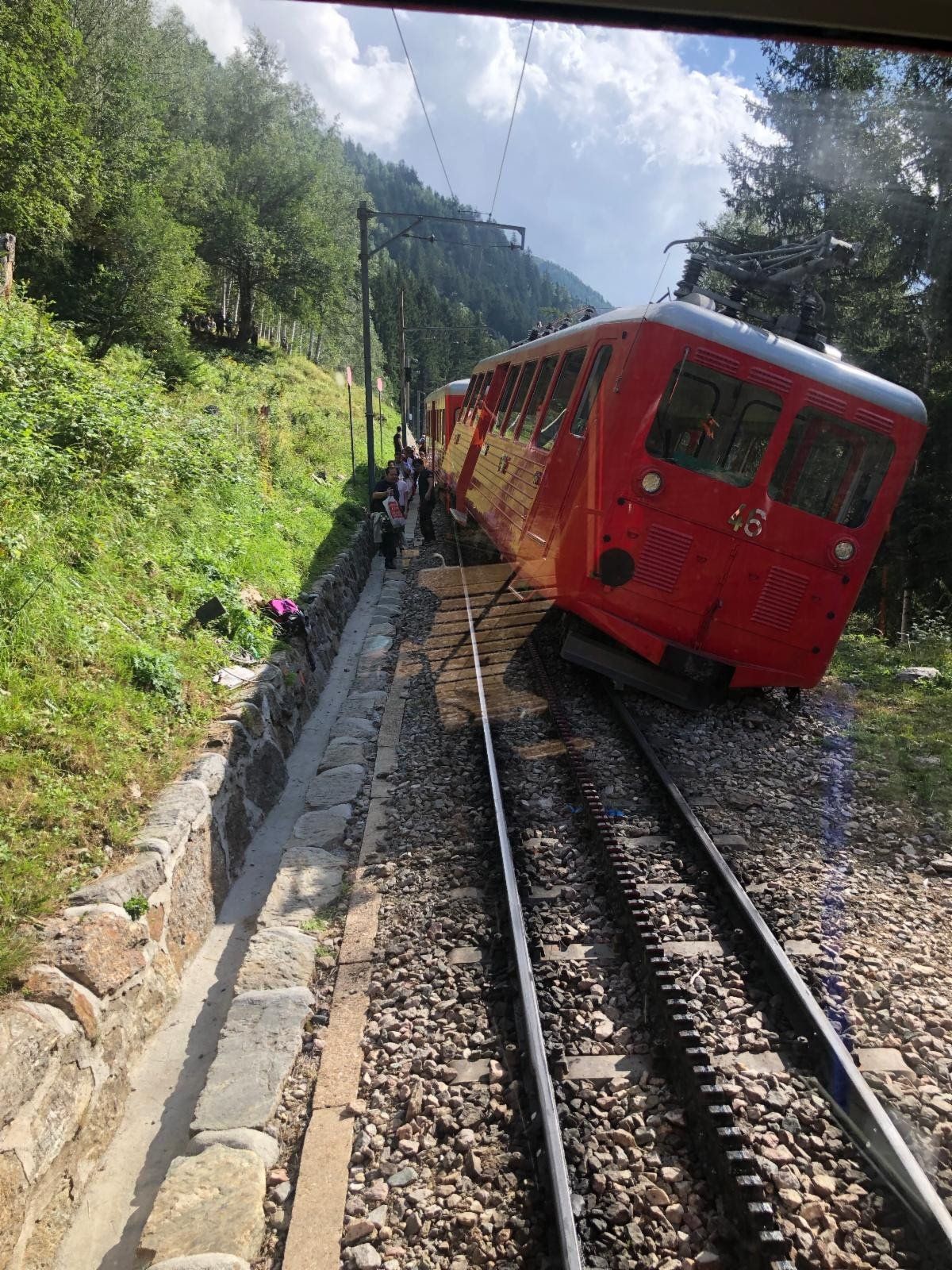 Red funicular train ascending a steep track on a mountain, with trees and a stone wall alongside.