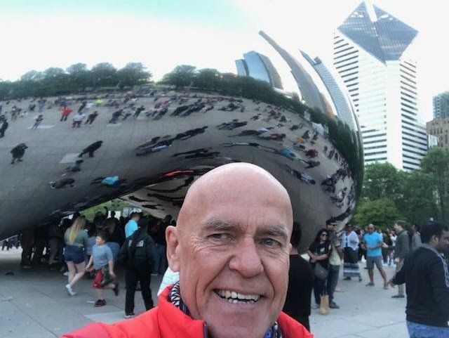 Man smiles in front of Chicago's Cloud Gate sculpture. Red jacket, buildings, and crowd visible.