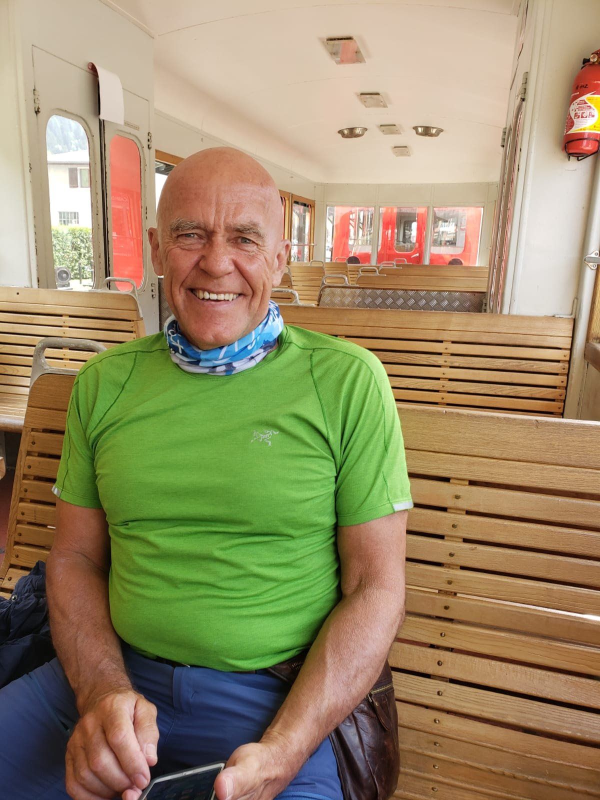 Bald man in green shirt smiles while sitting on a wooden bench inside a train car.