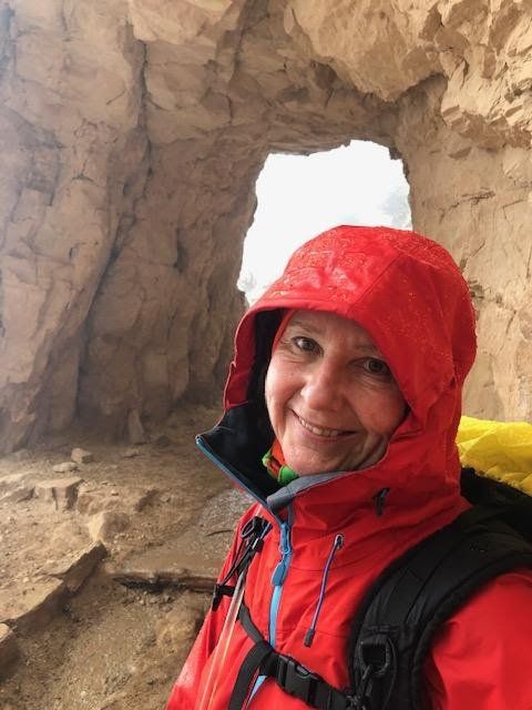Woman in red jacket smiles in a stone opening. Hiking with backpack.