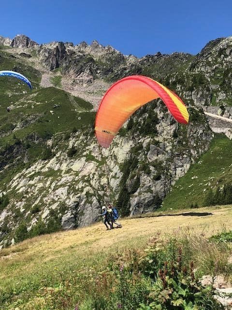 Person launching a red and yellow paraglider from a grassy mountainside, another paraglider in the distance.