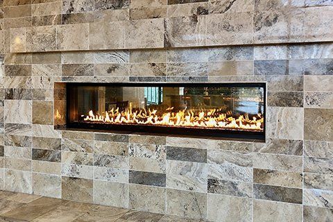 A modern fireplace with a long flame behind glass, set in a stone-tiled wall.