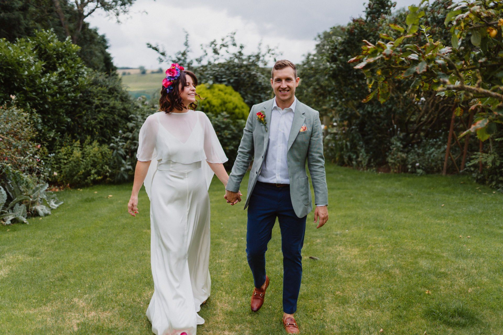 A bride and groom are walking through a grassy field holding hands.