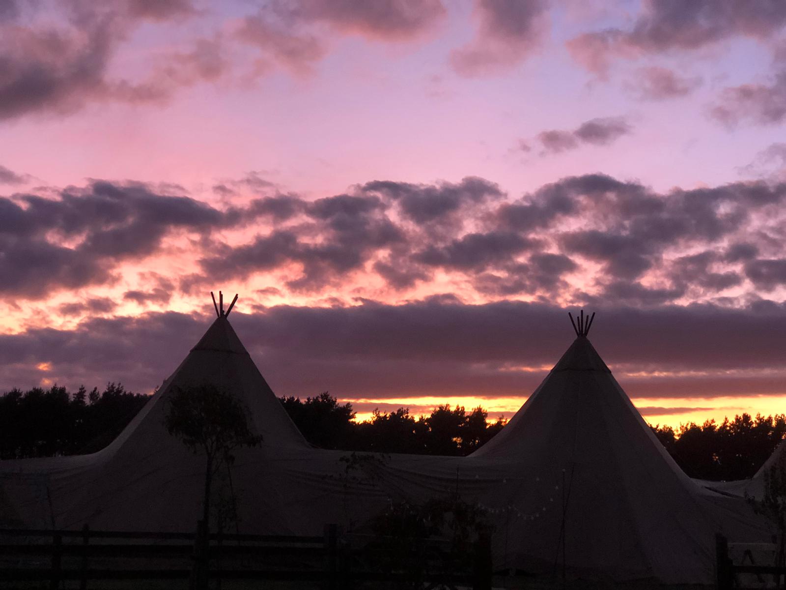 Two teepees are silhouetted against a sunset sky