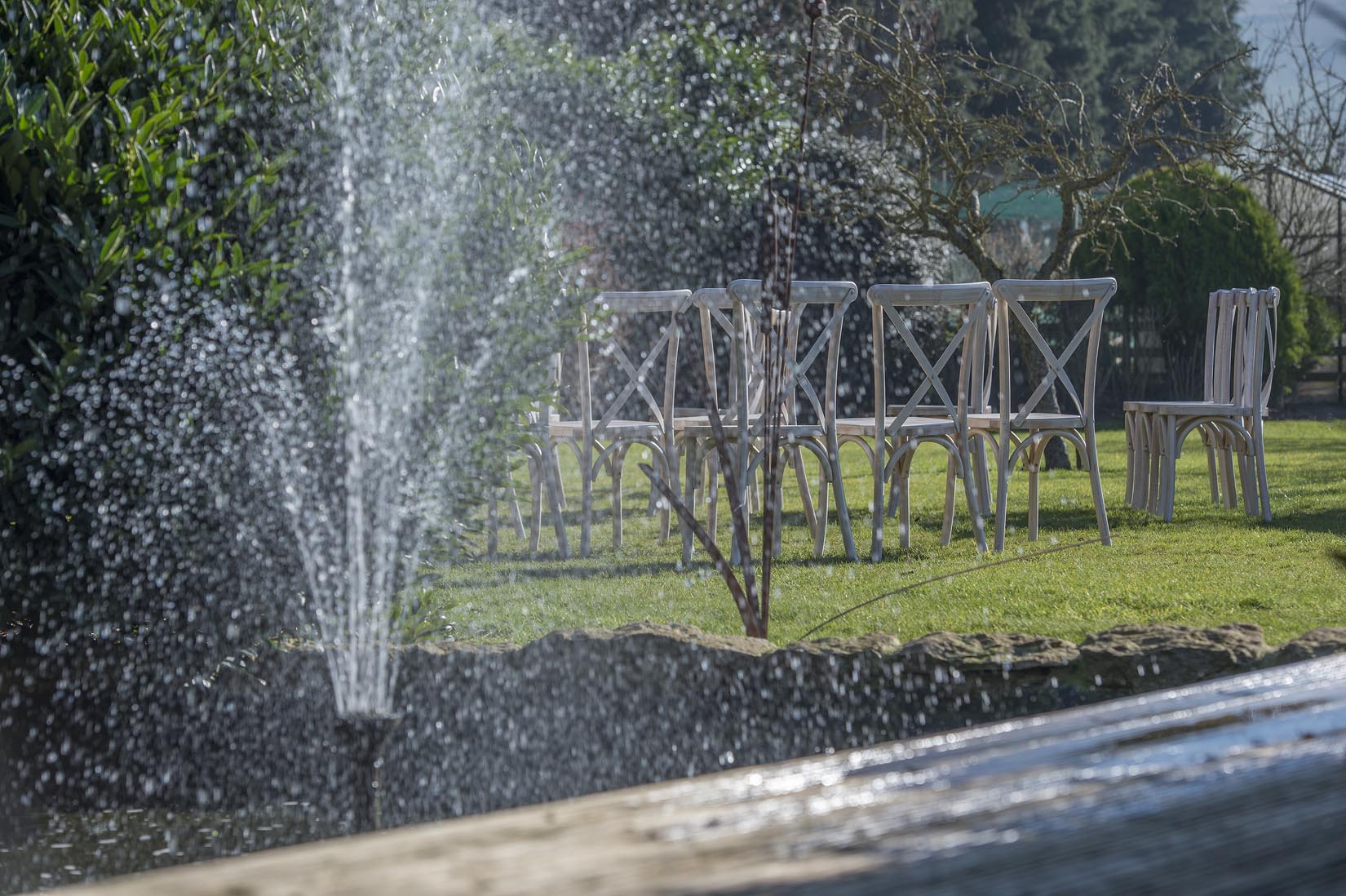 A fountain is spraying water over a row of chairs in a park.