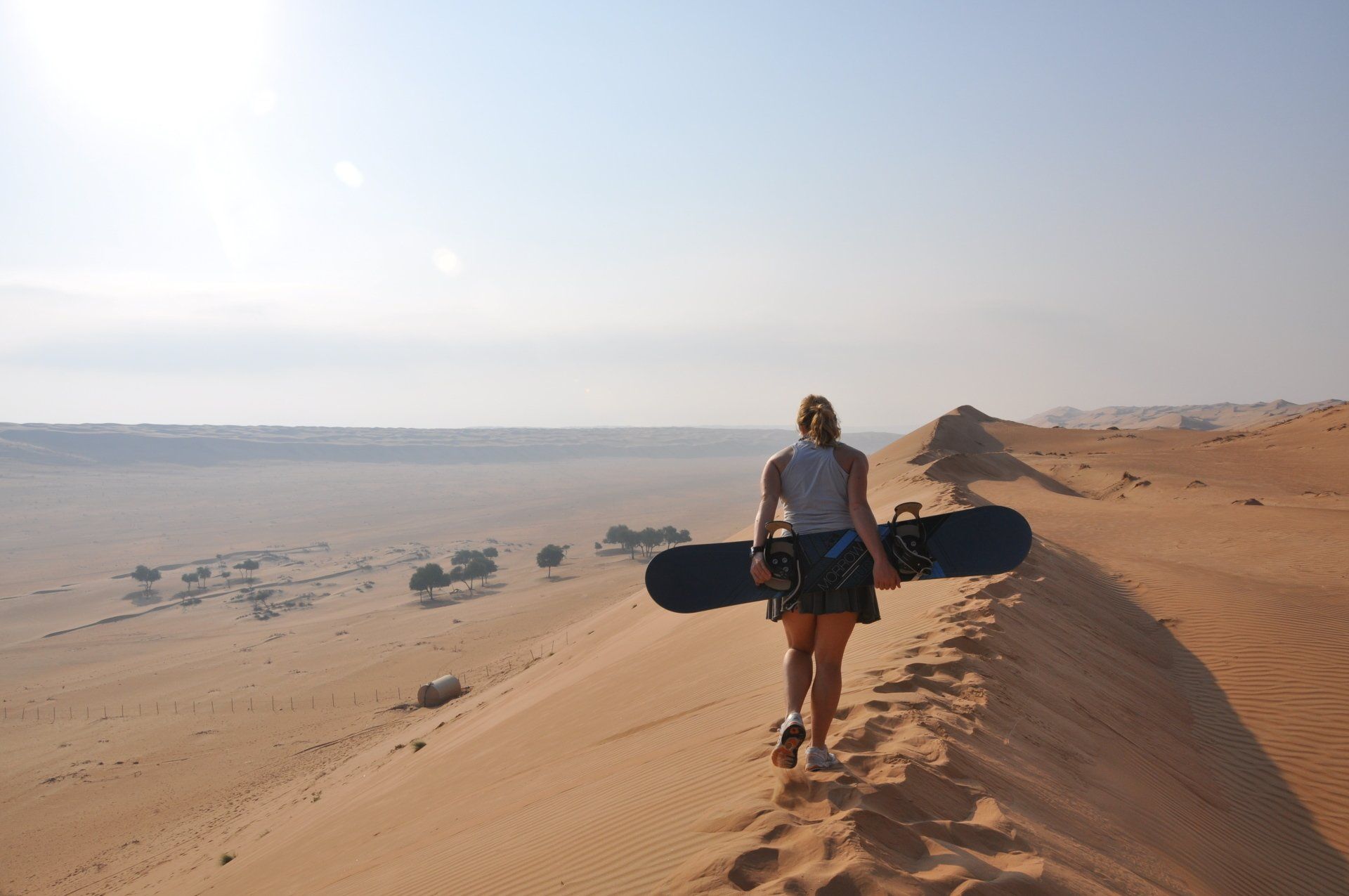 Person carrying a snowboard walks along a sand dune in a desert at sunrise.