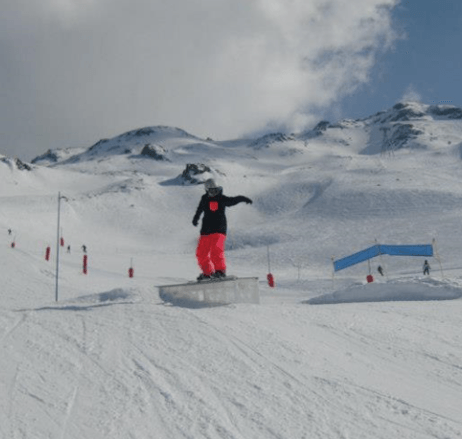 Snowboarder in black jacket and red pants balancing on a rail in a snowy mountain park.