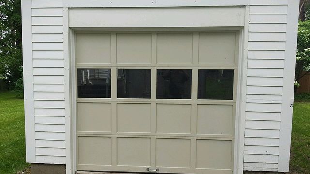 Tan garage door with four window panes centered in a white-sided building with green grass.
