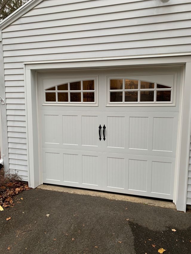 White garage door with arched window panels and black hardware, set in a light gray house.