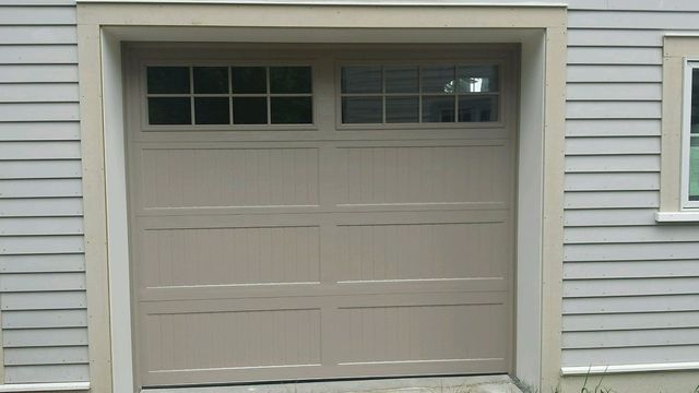 Beige garage door with square-pane windows, tan trim, and gray siding.