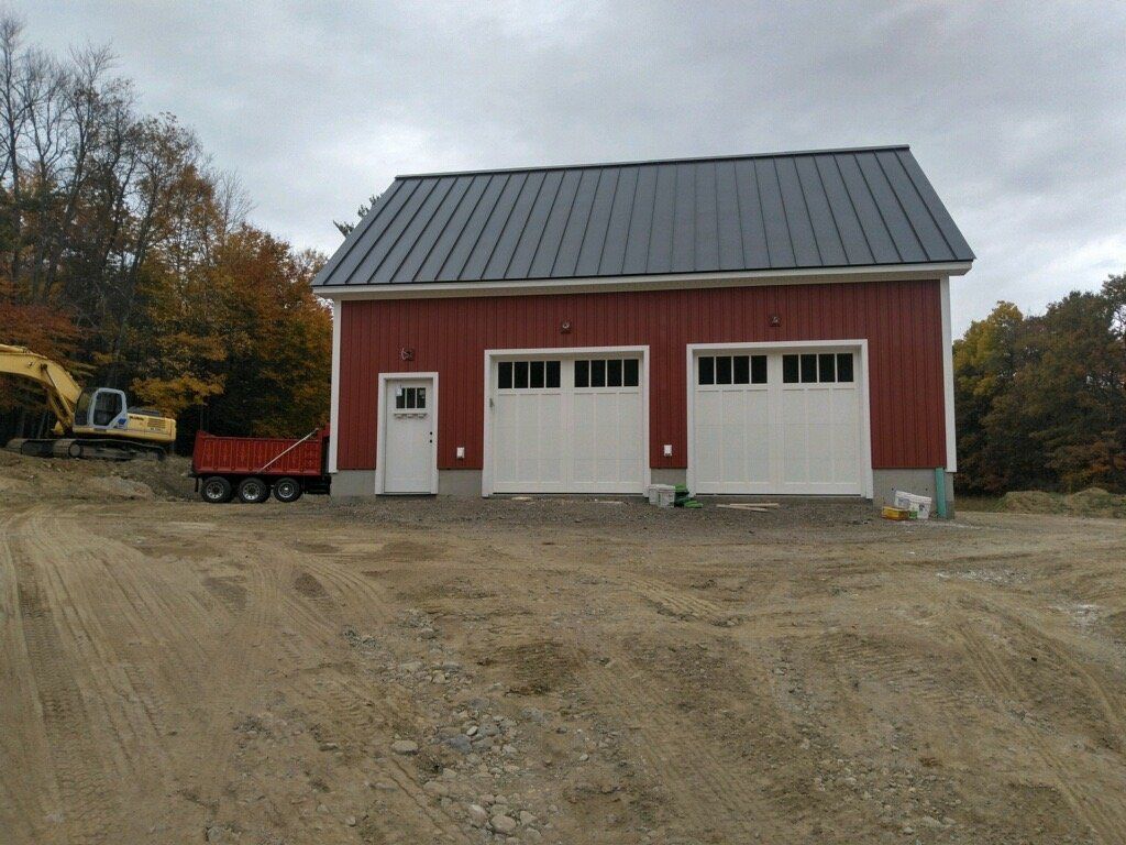 Red garage with two white doors, one side door, dark roof, dirt lot, excavator.