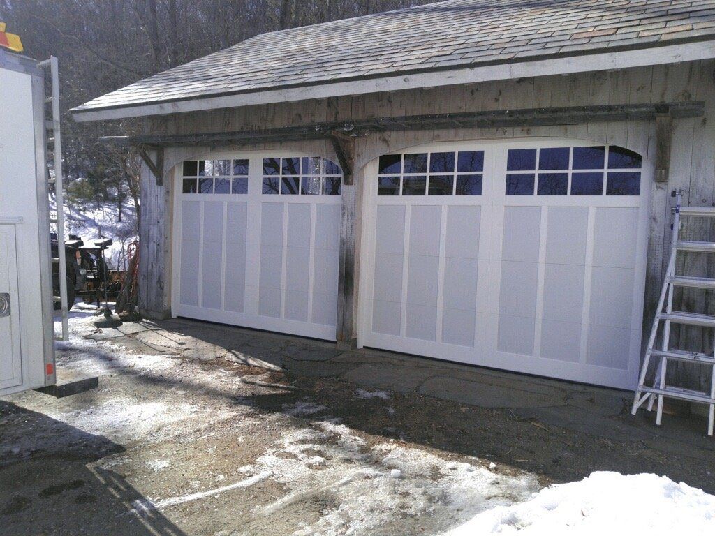 Two white garage doors with glass windows under a weathered roof, surrounded by snow and a ladder.