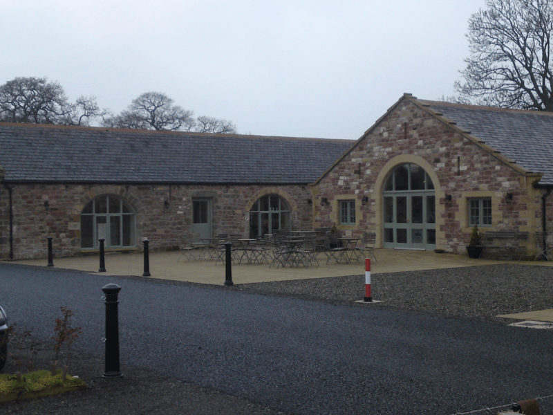 Stone building with arched windows and wooden doors beside a paved drive on an overcast day