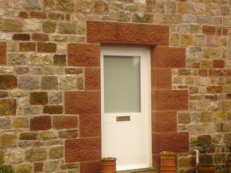 White door in a stone wall, framed by red brick, with a small potted plant below.