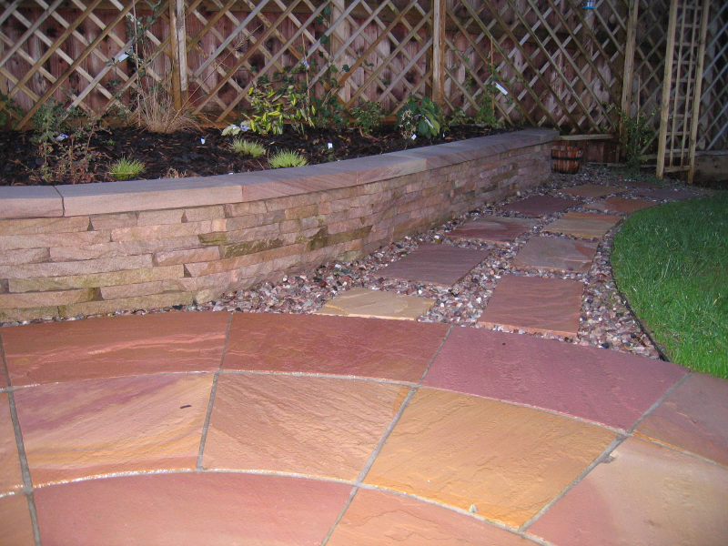 Curved terracotta patio tiles beside a stone retaining wall and lattice fence, with a strip of grass and gravel path