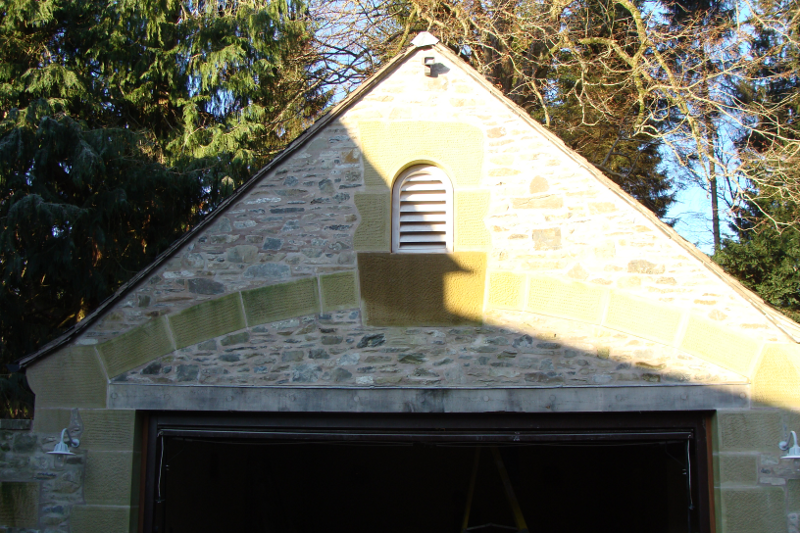 Stone garage gable with yellow siding and white vent in sunlight
